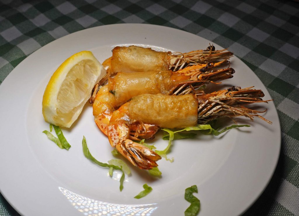 Drei frittierte Garnelen, Zitronenscheibe, Salatstreifen auf weißem Teller auf grün-weißem Tuch.