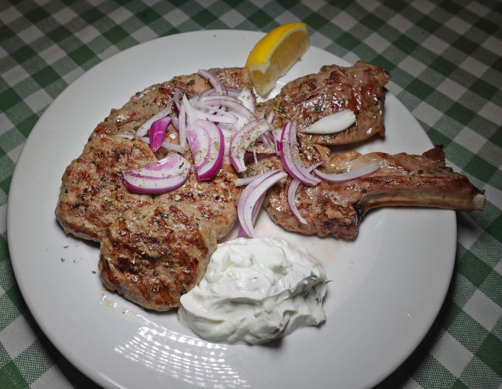 Gegrillte Koteletts mit roten Zwiebeln, Zitronenspalte und Tzatziki auf einem weißen Teller.