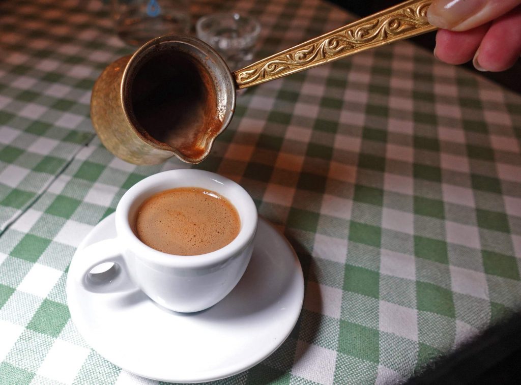 Eine Hand gießt Kaffee aus einem verzierten Kännchen in eine weiße Tasse auf einem karierten Tischtuch.